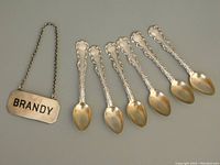 Overview of six small sterling silver coffee spoons with scalloped handle details and Brandy decanter tag with chain.