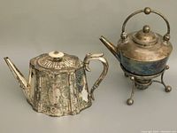 Photo showing both antique silver tea pots side by side, one octagonal with detailed engraving and the other on a stand with round ball design and base burner.