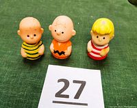 Front view of three vintage Charlie Brown Peanuts finger puppets (Charlie Brown, Schroeder, Lucy) on green cloth background with lot number card.