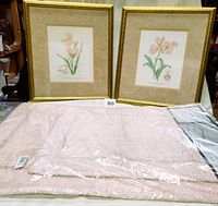Two gold framed floral pictures with pink matting displayed behind two pink bath mats in original packaging on a table.