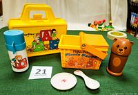 Front view of vintage Fisher-Price picnic basket set including brown bear thermos, white plate, white spoon, yellow picnic basket, blue ABC lunchbox and thermos.