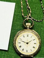 Close-up of lady’s silver fob watch showing white enamel dial with Roman numerals, gold decoration, and ridged crown.