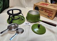 Fondue set pieces displayed separately: handled fondue pot with lid, black metal stand with tray, gel fuel/votive candle holder and snuffer, original packaging box in background.