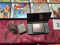 Black DS Lite open next to AC adapter with four games partially visible above