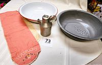 Vintage Spartan pewter creamer, gray speckled enamelware basin, white enamelware basin with red rim, and linen embroidered teaspoon holder laid on table