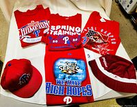 Lot laid out on white surface showing four red Phillies short-sleeve shirts and two caps. Shirts have graphics of Bryce Harper, 2009 World Series, 2009 Champion, and Spring Training 2020. Two caps include bucket hat with Phillies Chevrolet logos and fitted maroon Phillies cap.