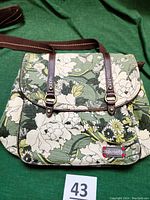 Front view of olive green Sakroots women's backpack with floral canvas pattern and two antique brass buckle straps on flap, showing front zippered pocket and Sakroots label.