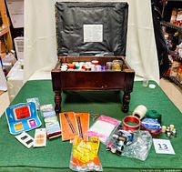 Photo of vintage wooden sewing chest with open lid revealing multiple spools of thread inside and sewing notions arranged in front on green fabric.