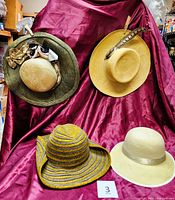 Four hats displayed on a burgundy fabric backdrop showing wide brim styles and various designs including feathers and ribbons.