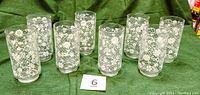 Eight vintage water glasses arranged in two rows on green cloth, showing white embossed flowers and lattice design on clear glass.