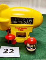 Front view of the yellow Good Weeble Blimp showing 'HAPPY BIRTHDAY' message on digital sign and the Tumblin Weeble pilot figure in front.