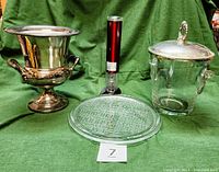 Full view of four items: silver-plated wine chiller on left, red Wolfgang Puck wine opener in back center, clear glass serving plate embossed with cheese names front center, clear glass ice bucket with silver-plated lid and handles on right.