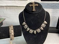 Photo of all jewelry items displayed together: cross pendant on stand, necklace draped on bust, and bracelet on a velvet holder.