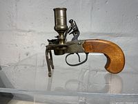 Side view of pistol form candlestick lighter showing wooden handle, brass and metal construction, and candlestick holder on top.
