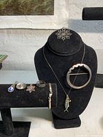 Group photo of sterling silver jewelry including pendants, rings, bracelet, brooch, earrings, necklace with fish-shaped pendant. Displayed on black jewelry stands and bracelet bars.