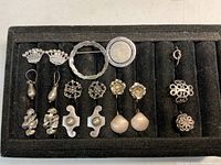 Top view of vintage sterling silver jewelry neatly arranged on black display with 7 pairs of earrings including crown shapes, teardrops, floral studs, shell drop earrings, two brooches and three rings.