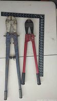 Photo showing two bolt cutters, a larger blue one and a smaller red one on a white surface with a measurement square for scale.
