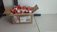 Open cardboard box filled with numerous small retail spark plug boxes, mostly Motorcraft brand with orange and white color scheme, placed on white table.