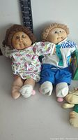 Two Cabbage Patch Kids dolls side by side, wearing pajamas and pants/shirts, showing soft fabric bodies with plastic faces and yarn hair, with measuring scale next to dolls.