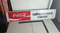 Red and white Coca Cola metal sign showing the full 'Enjoy Coca Cola refreshment center' text laid flat on a table with measuring rulers nearby.