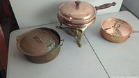 Photo of three copper kitchenware items displayed on a table: a large round pot with lid, an oval copper pan with lid, and a brass-copper chafing dish with legs and dome lid.