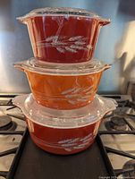 Stacked three Pyrex bowls in graduated sizes with wheat pattern design and lids, side view showing colors from deep red to orange gradient.