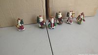 Photo of six Santa Claus ornaments in varied poses displayed in front of two large cardboard boxes labeled 'Galleria Los Angeles Made in China'.