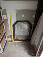 Black metal framed skid steer door with clear glass window propped vertically against a wall, a yellow measuring stick next to it for scale.