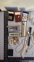 Photo showing assorted watches, pearl necklaces, pens, costume jewelry pieces including a brooch and gold-tone ring in display boxes.