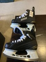 Pair of Bauer boys' ice hockey skates with white Tuuk blade holders and blades resting on a dark wood surface, showing side and top views.