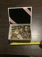 Box with pink ribbon detail open showing a collection of foreign coins inside, measuring tape alongside for scale.