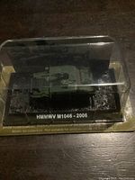 Front and side view of green diecast HMMWV M1046 military vehicle model with clear plastic display cover on black base with label and text.