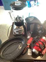 Photo showing Oster blender with glass jar, black electric kettle, large oval roasting pan, and red electric mixer with measured cups and cutlery nearby.