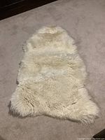 Image shows a natural lamb skin rug laid out on beige carpet floor, off-white cream color with fluffy wool texture visible.
