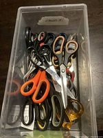 Top view of various scissors placed in a transparent plastic container showing different handle colors and blade sizes.