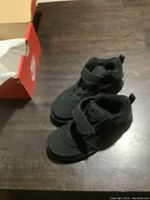 Pair of black baby Nike shoes sitting on a dark wood floor next to an open red Nike shoebox.