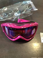 Pink Smith snow goggles shown from front angle with reflective tinted lens and black adjustable strap; placed on a surface with a clear plastic bag nearby