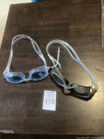 Two pairs of swimmers goggles displayed side by side on a wooden surface, one with clear blue lenses and one with black lenses, both with adjustable white straps.
