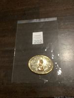 Photo of the oval wolf belt buckle inside a clear plastic bag on a dark surface showing its detailed engraving and central wolf figure.