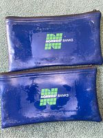 Two blue Norwest Banks vinyl zip pouches placed flat to show front side with logo. The surface has wear, scratches, and tape residue marks.
