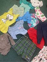 Photo showing assortment of baby clothes including yellow Snoopy tank, light blue piece, plaid shorts, and various pants and pajamas.