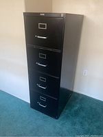 Front and angled view of the 4 drawer black metal filing cabinet against a white wall on green carpet.