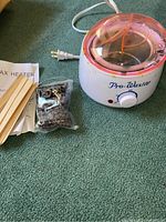 Photo showing wax heater unit with clear pink lid, instruction manual, bag of dark wax pellets, and wooden sticks on carpeted floor.