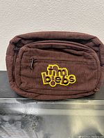 Front view of brown corduroy fanny pack with yellow and brown embroidered 'tim biebes' logo on front zippered pocket.