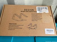 Photo of the cardboard box for the Design LS03 laptop stand showing product branding, usage instructions, company contact info, and an illustration of how parts A and B connect.
