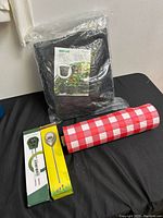Photo showing new black fabric grow bags sealed in plastic packaging, soil thermometer in green packaging, soil moisture meter in yellow packaging, and rolled red and white checkered tablecloth.