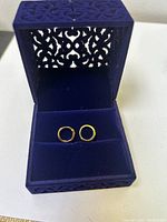 Pair of small gold plated sterling silver hoop earrings inside a dark blue decorative jewelry box with intricate cut-out pattern.