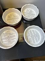 Four white scalloped edge ceramic mini pie dishes displayed on glass surface; each dish has a baking recipe printed inside the bottom surface