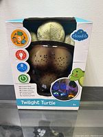 Front view of the boxed Twilight Turtle night light showing plush turtle and star projection dome with icons for 3 light modes, 8 colors, and timer.