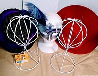 Photo showing 2 white metal wireframe hat stands, one black woven hat with purple scarf and blue feathers, wide-brimmed red hat, and white Styrofoam head wearing a blue and silver feathered mask.